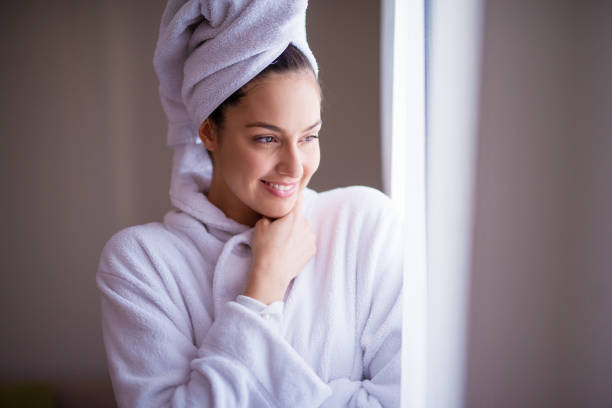 joyfull hermosa joven mujer en una bata con una toalla alrededor de su pelo es sonriente y sensación fresca después de la ducha mientras mira por la ventana y sentir acogedor. - bathtub women bathroom relaxation fotografías e imágenes de stock