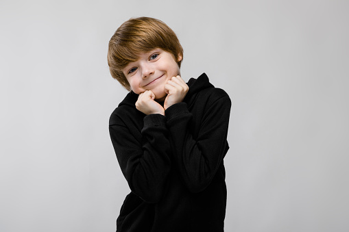 Cute caucasian little boy in black sweater looking in camera smiling sweet with hands near his face
