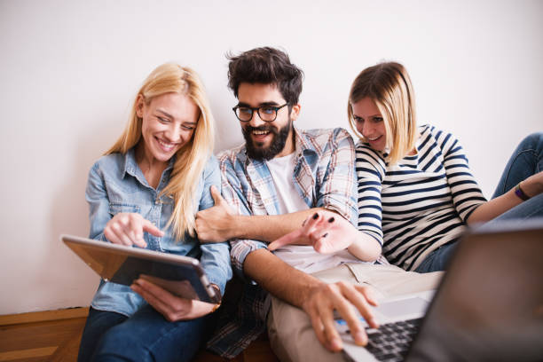 istock Group of young playful coworkers laughing on internet content from a tablet while sitting on the floor leaning against the wall. 944894314
