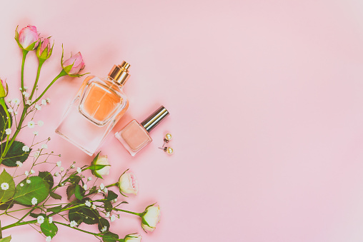 Flat lay of female cosmetics products and accessories. A bottle of perfume, nude nail polish, pearl earings and roses over pink background. Copy space.
