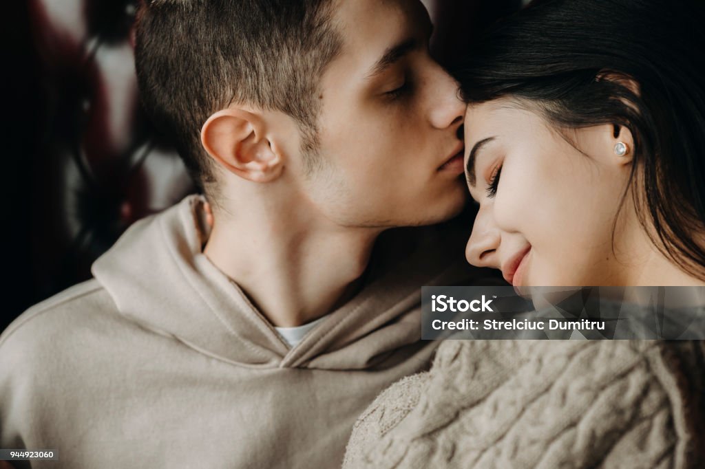 Close up portrait of a lovely young couple sitting on a leather chair while boy is kissing his girlfriend on forehead. Below Stock Photo