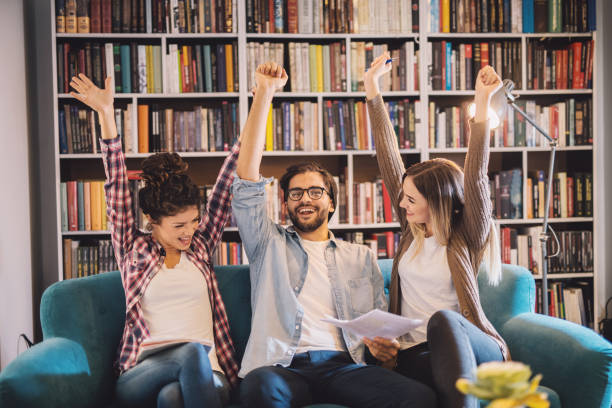 grupa uroczych podekscytowanych studentów świętujących zakończony egzamin siedząc na kanapie w bibliotece. - book sofa women bookshelf zdjęcia i obrazy z banku zdjęć