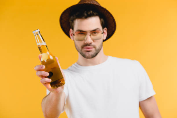 handsome young man in hat and sunglasses holding bottle with summer drink and looking at camera isolated on yellow