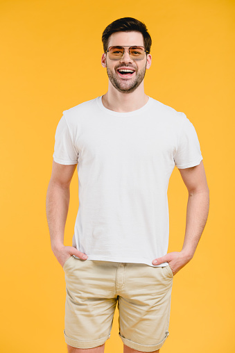 handsome bearded young man in sunglasses standing with hands in pockets and smiling at camera isolated on yellow
