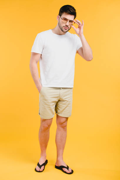 istock handsome young man in shorts and flip-flops adjusting sunglasses and looking at camera isolated on yellow 944974724