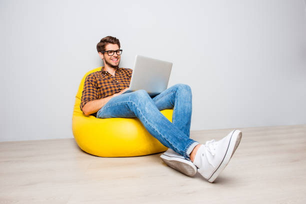 행복 한 젊은 사람이 노란색 pouf에 앉아서 노트북을 사용 하 여 - elegance yellow glasses eyewear 뉴스 사진 이미지