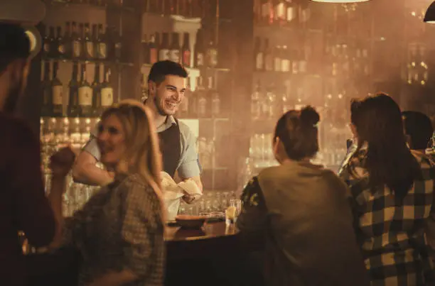 Young happy waiter cleaning glasses and communicating with his customers in a bar.