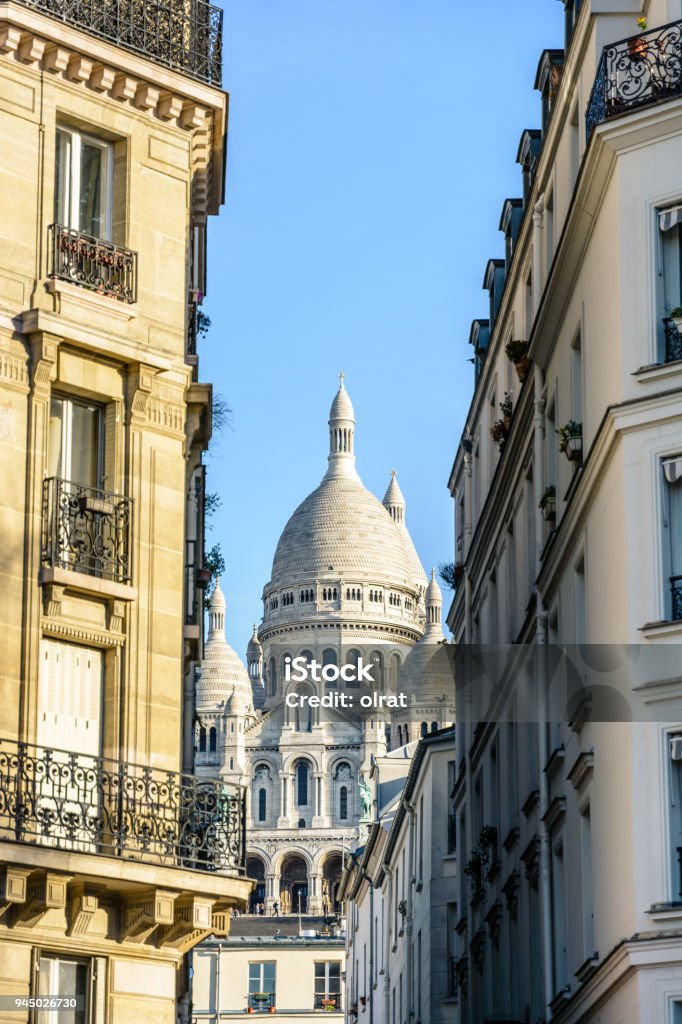 View of the facade and dome of the Basilica of the Sacred Heart in Paris through a narrow street between typical buildings under a clear blue sky. Apartment Stock Photo