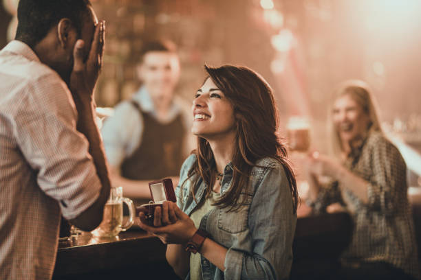 Happy woman proposing to her boyfriend in a bar. Young happy woman proposing her black boyfriend during night out in a pub. There are people in the background. engagement stock pictures, royalty-free photos & images
