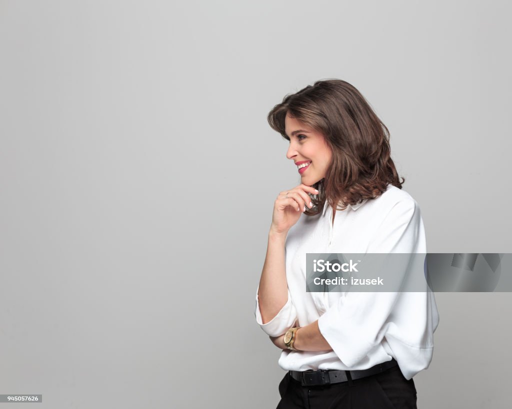 Side view of beautiful young businesswoman against grey background Portrait of confident young businesswoman standing with hand on chin and smiling against grey background. Side View Stock Photo