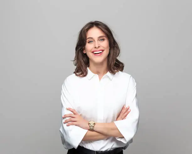 Portrait of confident young businesswoman standing with hand crossed and smiling against grey background.