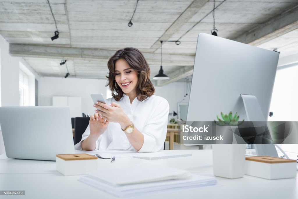 Smiling businesswoman making a phone call in office Cheerful young beautiful woman typing on mobile phone while sitting at her working place. Smiling businesswoman making a phone call in office. Businesswoman Stock Photo