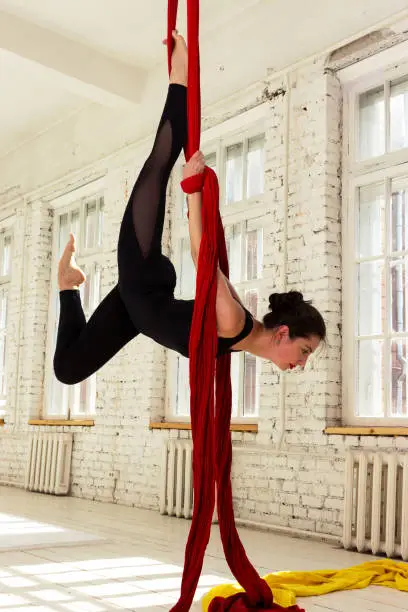 Beautiful aerialist girl doing acrobatic and flexible tricks on red aerial silks (tissues) on white brick and white wall on background
