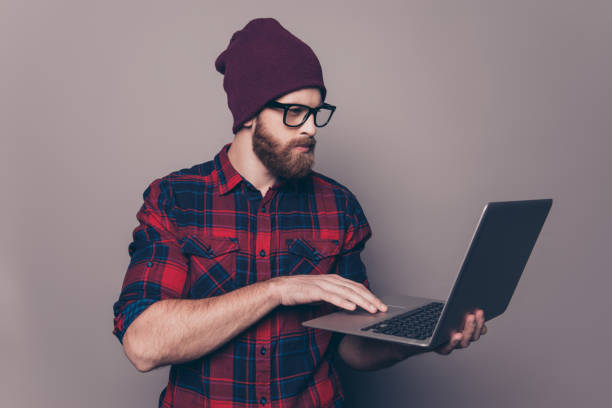 istock Smiling handsome young man in cap and glasses holding modern laptop 945075738