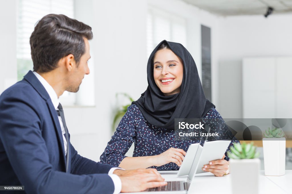 Muslim businesswoman in discussion with colleague Smiling muslim businesswoman holding digital tablet and discussing work with businessman in office. Businesspeople discussing new project. Business Stock Photo