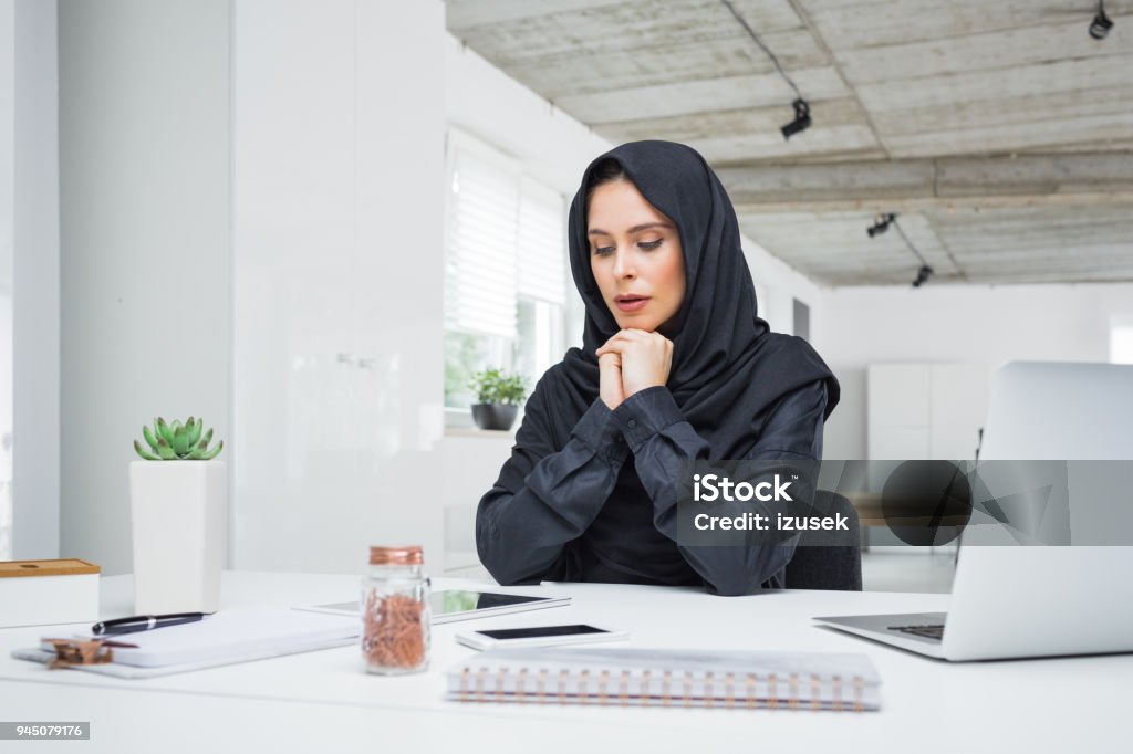 Worried muslim business woman working in office Worried business woman working on her laptop in the office. Muslim female in head scarf sitting at her desk. Adult Stock Photo