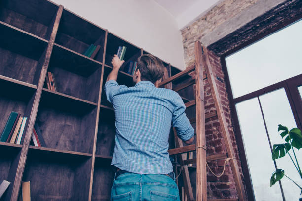 istock Rear view of man standing on stepladder and searching book in library 945086092