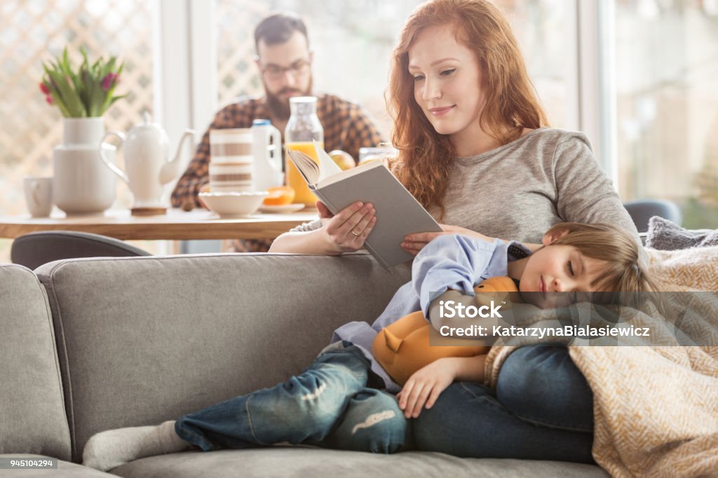 Son lying on mother's laps Sleeping son lying on mother's laps while she is reading a book and father eating in the background Family Stock Photo