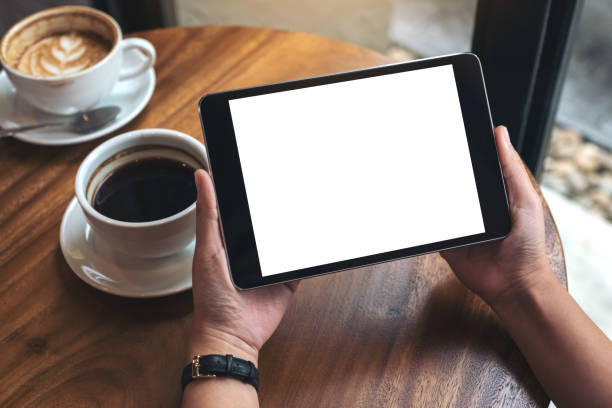 istock Mockup image of hands holding black tablet pc with white blank screen and coffee cups on table 945120070