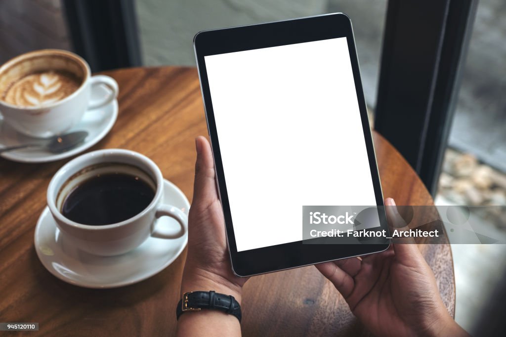 Image de la maquette des mains tenant noir tablet pc avec écran blanc blanc et tasses à café sur la table - Photo de Tablette numérique libre de droits