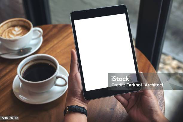 Mockup Image Of Hands Holding Black Tablet Pc With White Blank Screen And Coffee Cups On Table Stock Photo - Download Image Now