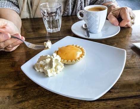 Woman's hand holding fork with bite of Lemon tart and clotted cream -coffee cup setting on table beside plate