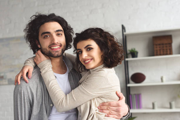 istock smiling girlfriend hugging boyfriend and they looking at camera at home 945141764