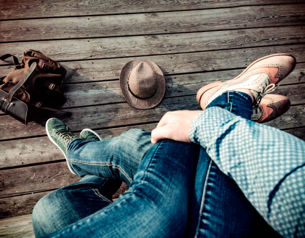Couple is sitting embracing their legs on wooden floor in sneakers and boots and blue jeans on the outdoors. On the floor lie a straw hat and a backpack. Concept of meeting after a long separation. pareja está sentada abrazando sus piernas en el piso de madera en zapatillas y botas y pantalones vaqueros azules en el exterior. en el piso se encuentran un sombrero de paja y una mochila. concepto de reunión después de una larga separación