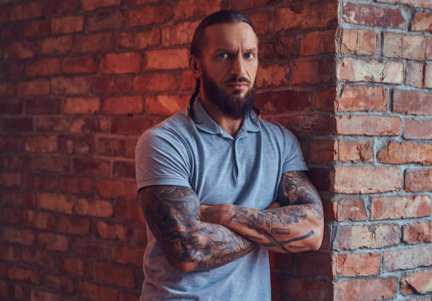 スタイリッシュな散髪とひげ、グレーの t シャツに tattoed ハンサムな男性は、ロフトのインテリアをお部屋をレンガの壁にもたれて立っています。 - tattoo t shirt brick wall portrait ストックフォトと画像