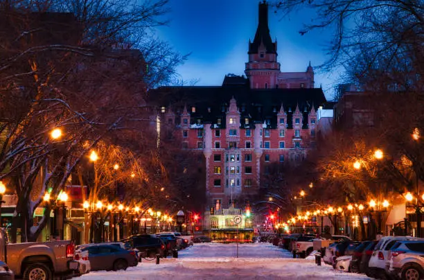 A snow cover street illuminated by street lights on both sides with a tradition Canadian style building at the end of the street with a glowing blue sky.