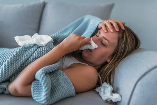 Sick Woman having Flu with Caught Cold. Sneezing into Tissue. Headache. Virus. Sick woman lying in bed with high fever. She is blowing nose. Photo of Young female Lying On Bed covered with a blanket  Infected With Allergy holding her head.