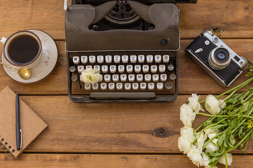 Writer flatlay. Dark gray retro typewriter with notepad, black pen, cup of black coffee, vintage film camera and white ranunculus over wooden background. Copy space.