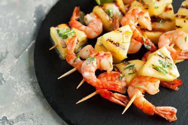 Grilled shrimp skewers with pineapple. Seafood, shellfish. Prawns skewers with herbs, pineapple, sweet chili and lime on black stone plate