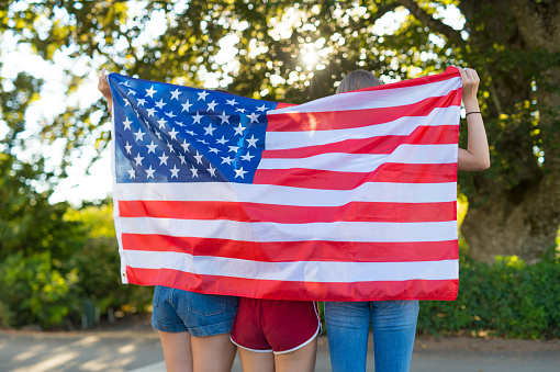 Three teenage girls pose with a large U.S.A. flag around them. They are standing on a sidewalk outside in front of a wooded area and holding it up behind them as they face away from the camera.