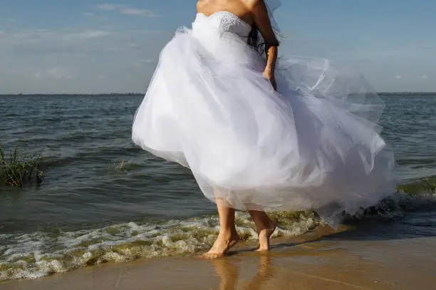 The bride on a sunny summer day runs barefoot on the surf line of the sea in a lush white dress. Girl in wedding dress running along the beach