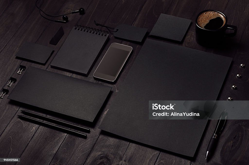 Black blank stationery set with phone, earphone, coffee on dark wood background, inclined. Template Stock Photo