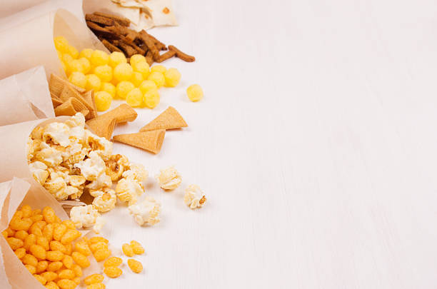 Golden colorful snacks - nachos, popcorn, croutons, chips in craft paper cone as border on white wood table with copy space, closeup, blur.