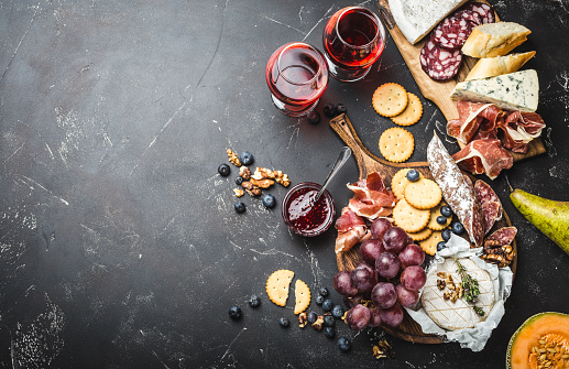 Red wine glasses, wooden boards, cold meat, cheese, fruit, dip. Stone background. Space for text. Delicatessen plate. Mix of different snacks/appetizers. Top view. Fruit, cheese, meat, wine. Party