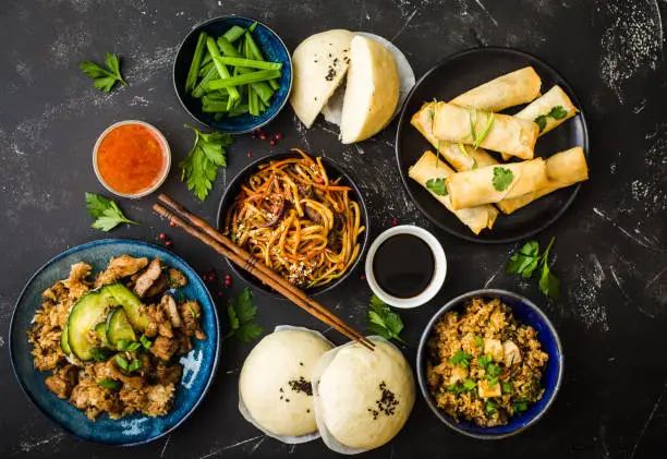Asian assorted food set, dark rustic stone background. Chinese dishes. Chinese stir-fry noodles, asian rice with meat, dim sum, fried spring rolls, steamed Chinese buns. Top view. Asian stile dinner