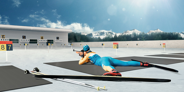Athletic woman at the biathlon competitions preparing to make a shot. Blue sky and snowy mountains in background. Female Biathlete has a gun and skis.