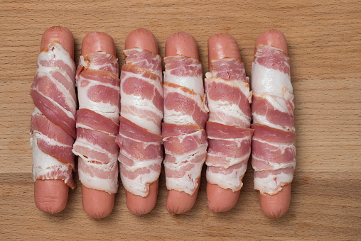 Preparation of raw sausages wrapped spirally in bacon on a wooden background. Top view.