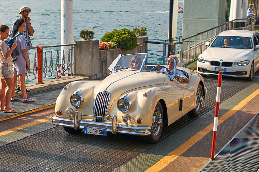 Bellagio, Italy: July 08 2017 - Happy man drives out his vintage beige Jaguar XK120, a sports cabriolet car manufactured circa 1950 in United Kingdom, from the ferry boat at lake Como.