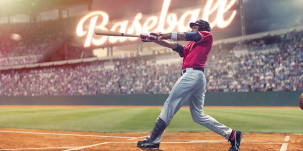 Professional Baseball Batter Striking Baseball During Night Game In Stadium Professional baseball player with baseball bat outstretched having just struck baseball during an outdoor baseball game. The batter is dressed in generic white and red baseball uniform with black safety helmet and stands in the batters box in front of spectators and a bright neon sign in a generic stadium. baseball player photos stock pictures, royalty-free photos & images