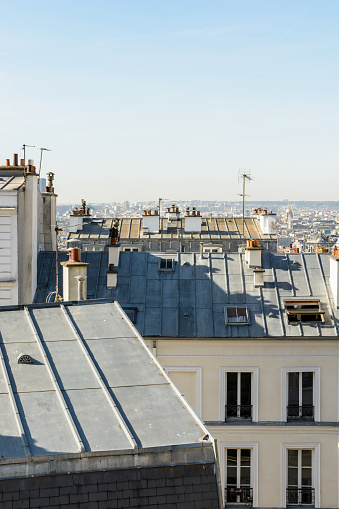 Open view over the rooftops of Montmartre district in Paris with chimneys, TV antennas, skylights and a typical zinc roofing in the foreground under a clear blue sky.