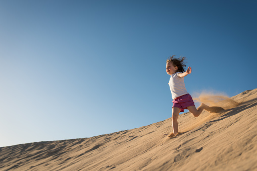 Little joyful girl running down sand dune on sunny day with clear blue sky