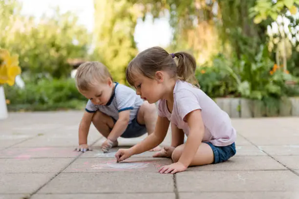 Brother and sister drawing together drawing with chalk outdoor on paving