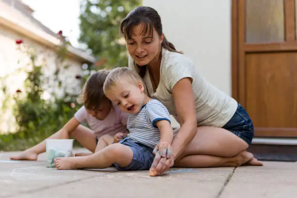 Mother is teaching her little son drawing with chalk in backyard and daughter is also drawing