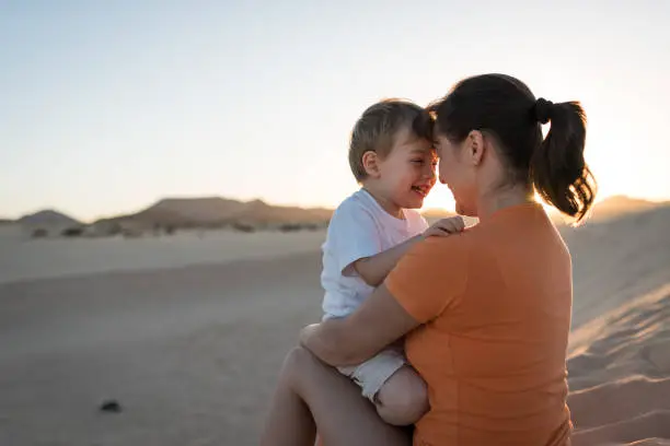 Happy little smilling boy hugging with his mother on sand dune in desert during sunset