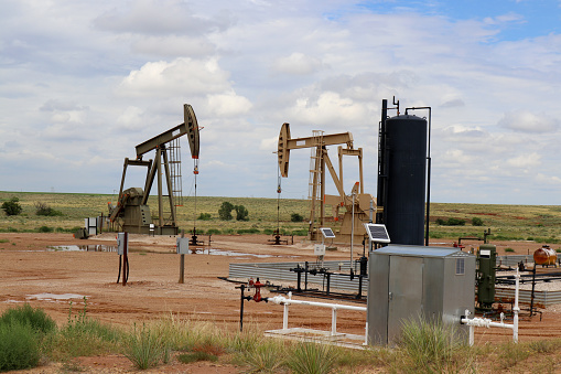 Two oil - gas well pump jack at drilling site with equipment and holding tanks on praries with low hill behind and blue cloudy sky