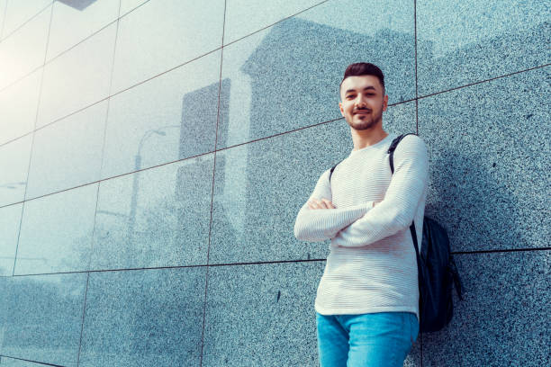 istock Arabian student with backpack standing by the wall outside. Young man waiting for groupmates. 945892068